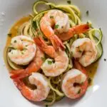 Creamy keto friendly dish made with garlic butter sauce and zoodles. Dish is plated on a white dish with shrimp and zoodles and parmesan